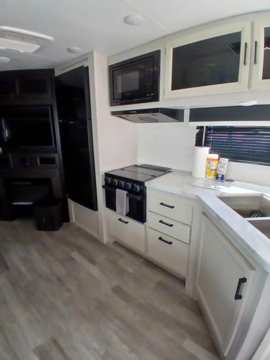 Inside an RV rental: a bright kitchen with black appliances, white cabinets, and wood-look flooring, leading to a cozy living area.. Jayco White Hawk 2023