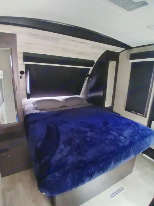 Cozy RV rental bedroom with a blue blanket, blackout blinds, and modern light wood-paneled walls. Perfect for your trip.. Jayco White Hawk 2023