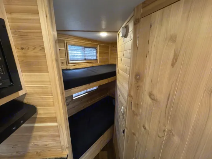 RV rental interior featuring cozy wood-paneled bunk beds with black mattresses and a small window, ideal for sleeping.. Ice Castle Fish Houses Ice Castle Fish Houses Trailer 2023
