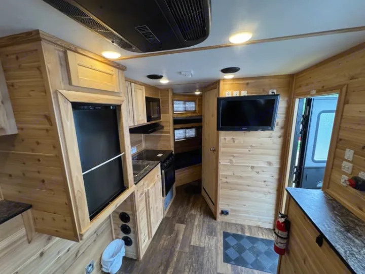 This rv rental interior features a full kitchen with a fridge & stove, bunk beds, and a TV mounted on wood-paneled walls.. Ice Castle Fish Houses Ice Castle Fish Houses Trailer 2023