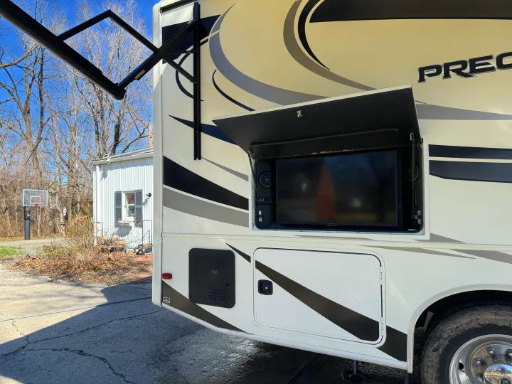 An RV rental showing an open outdoor entertainment compartment with a TV, speakers, and an extended awning arm overhead.. Jayco Precept 2021