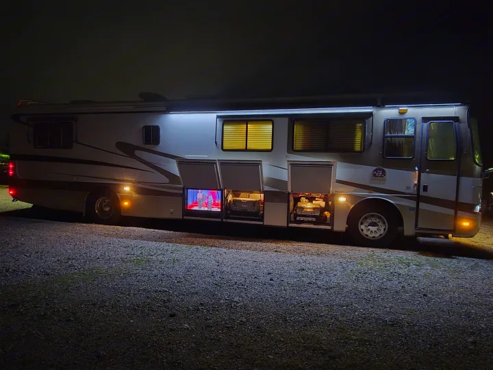 Nighttime RV rental setup: motorhome with outdoor TV, grill, and illuminated interior for a cozy camping experience.. Monaco Dynasty 2000
