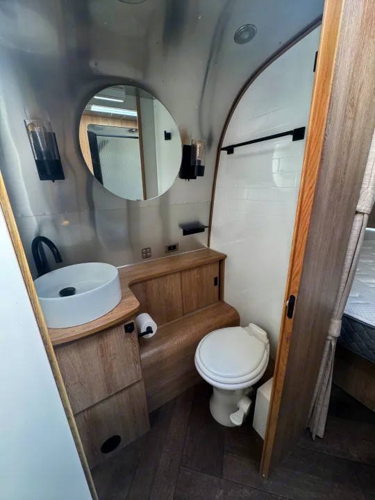 Sleek RV rental bathroom: vessel sink, toilet, subway tile shower, and wood cabinetry against curved metallic walls.. Airstream Pottery Barn 2023