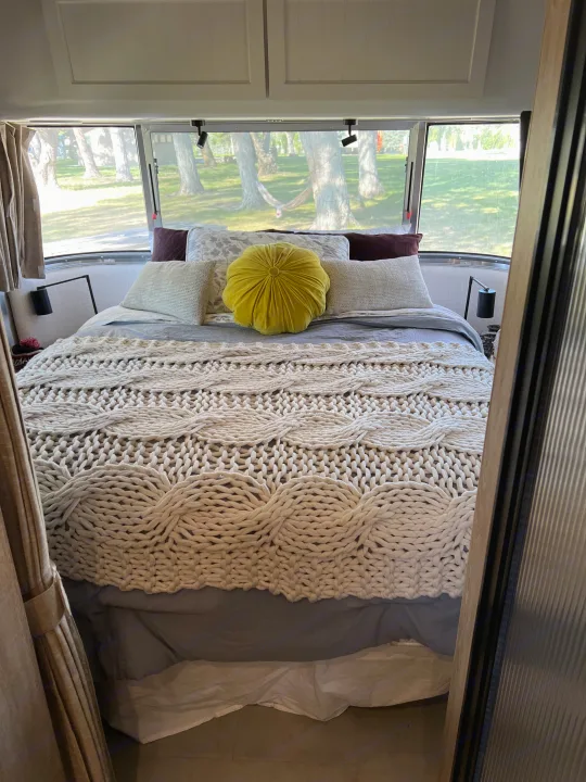 Cozy RV rental bedroom featuring a bed with a chunky knit blanket & yellow pillow, offering serene views through its windows.. Airstream Pottery Barn 2023