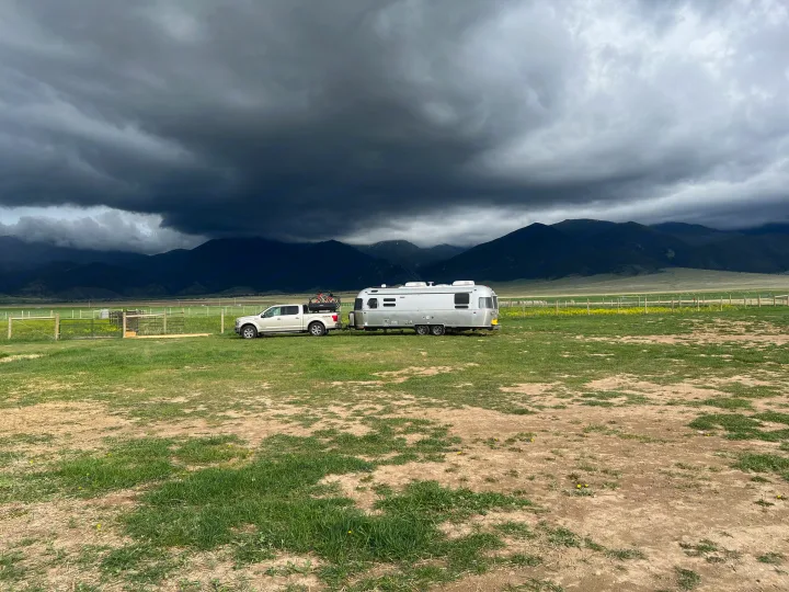 Stormy mountain views from your RV rental! A white pickup tows a silver Airstream in a grassy field under dark clouds.. Airstream Pottery Barn 2023