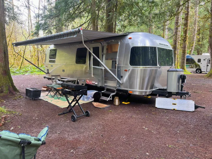 An Airstream rv rental parked in a mossy forest campsite with its awning extended, grill, and outdoor gear.. Airstream Pottery Barn 2023