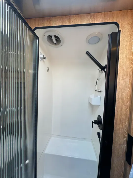 Clean, compact RV rental shower with clear reeded door, black fixtures, and ceiling vent/light. Perfect for travel.. Airstream Pottery Barn 2023