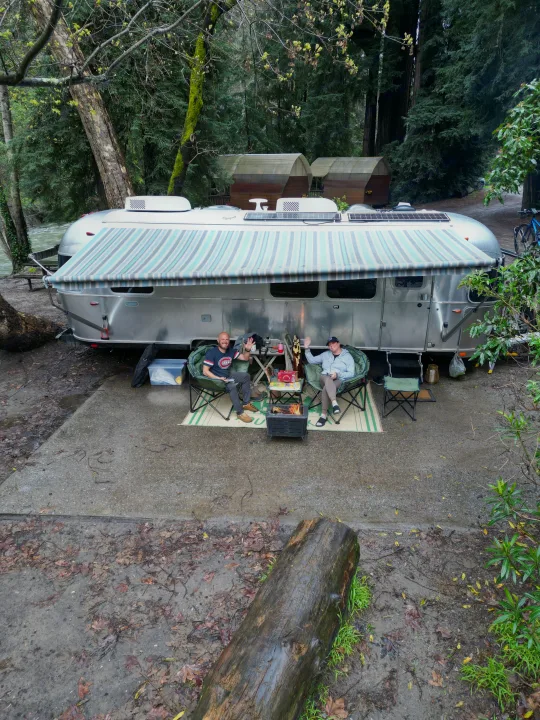 Two people waving from their rv rental Airstream setup, enjoying a campfire by the river in a wooded campground.. Airstream Pottery Barn 2023