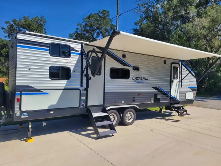 A grey and blue Catalina Legacy Edition travel trailer rv rental is parked on pavement, with its awning extended.. Coachmen catalina 2022