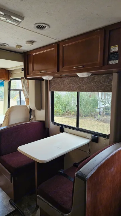 Interior of an RV rental showing a cozy dinette with burgundy cushions, wood cabinets, and an outdoor view.. Forest River Coachmen Pursuit 2014
