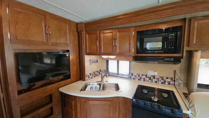 RV rental kitchen with wood cabinets, TV, microwave, double sink, stove, and tile backsplash on a light countertop.. Forest River Coachmen Pursuit 2014