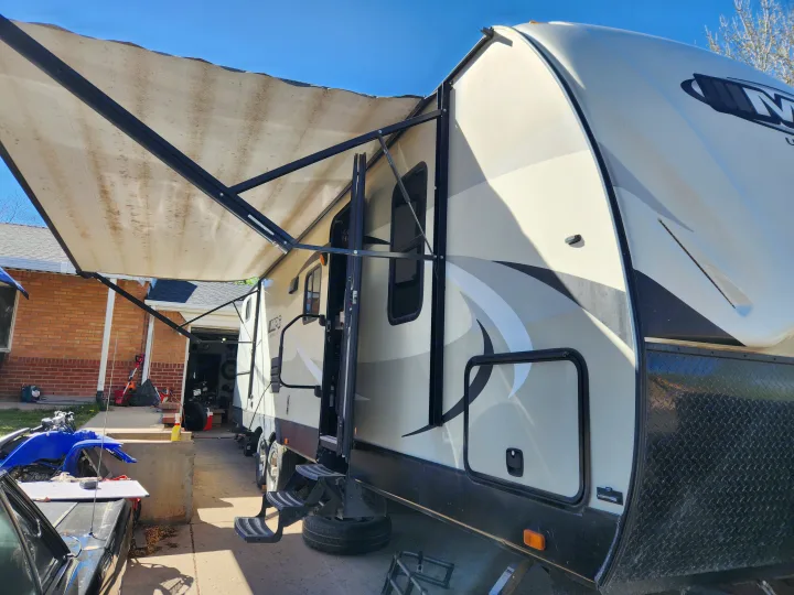 RV rental: A light-colored travel trailer with an extended, soiled awning, side graphics, and deployed steps, parked by a brick house.. Cruiser RV MPG Ultra Lite 2017