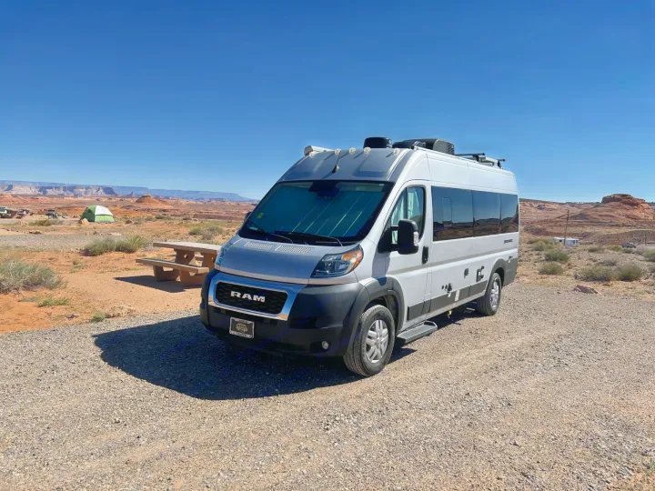 A silver rv rental camper van is parked at a sunny desert campsite with a picnic table and red rock mesas under a clear blue sky.. Thor Sequnce 2023