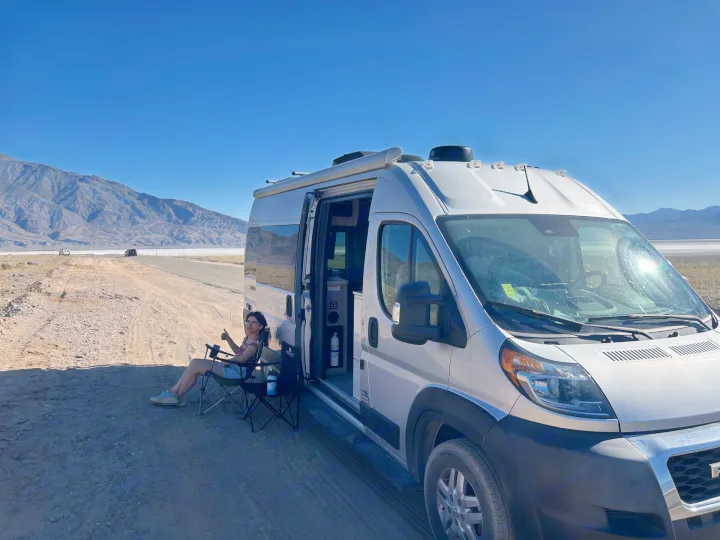 Woman gives a thumbs-up from her chair next to an RV rental camper van in a vast desert landscape with mountains.. Thor Sequnce 2023