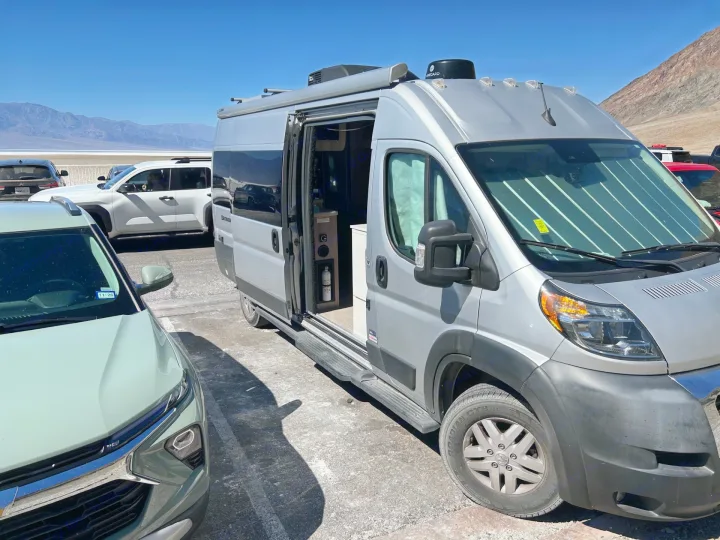 Silver RV rental parked in a sunny desert landscape with mountains in the background. Side door open, sunshade on windshield.. Thor Sequnce 2023