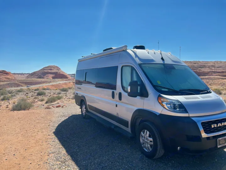 Silver rv rental camper van parked on a desert gravel road, with red rock hills under a bright blue sky.. Thor Sequnce 2023