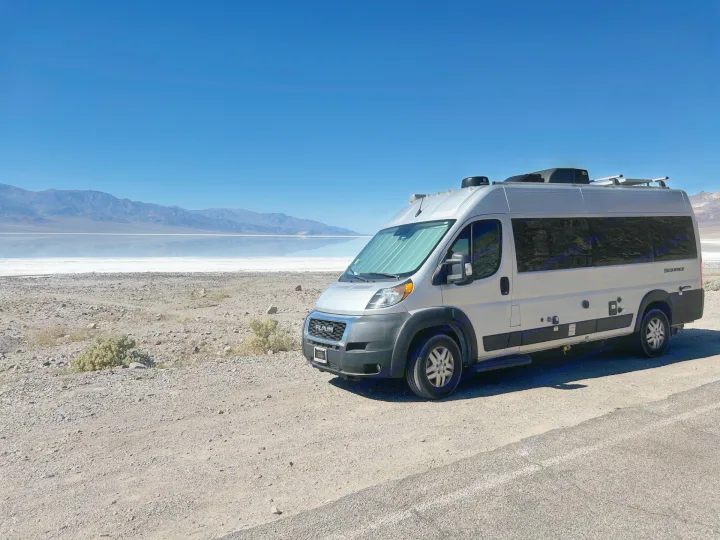 Silver RV rental camper van parked at Death Valley's reflective salt flats, with mountains under a clear blue sky.. Thor Sequnce 2023