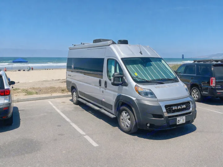Ram ProMaster RV rental parked at a sunny beach. Ocean, mountains, & people under a blue canopy in the background.. Thor Sequnce 2023