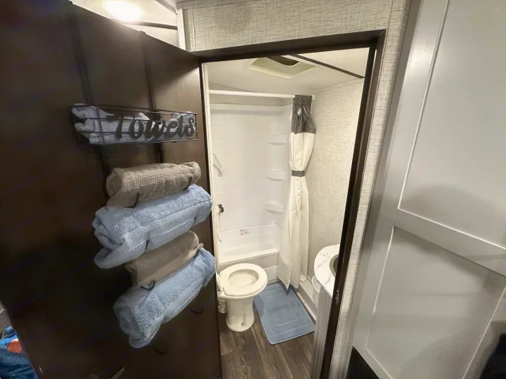 RV rental's compact bathroom showing a shower, toilet, blue bath mat, and neatly stacked towels on a dark wall rack.. Keystone RV Bullet Ultra Lite 2021