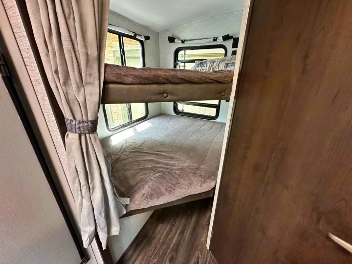Inside an RV rental, two bunk beds with soft blankets, windows, light curtains, and wood-look flooring create a cozy sleeping area.. Keystone RV Bullet Ultra Lite 2021