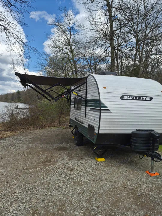 Sun Lite rv rental travel trailer with an extended awning, parked on gravel by a lake. Bare trees and a distant person on a dock.. Sunlite 16BH 2025