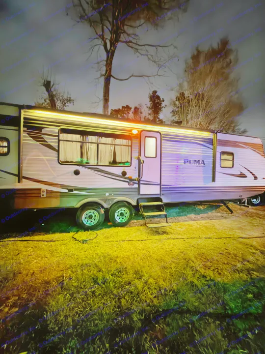 Nighttime rv rental: A "PUMA" travel trailer brightly lit with warm awning lights & purple hues, parked on grass beneath dark trees.. Forest River Palomino Puma 2018