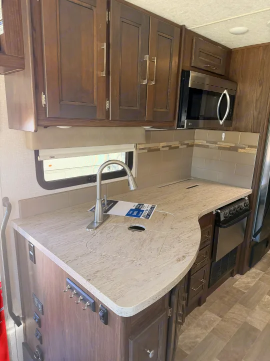 This RV rental kitchen has dark wood cabinets, a speckled light countertop with a sink, a microwave, and a small oven.. Coachmen Mirada 32SS 2019