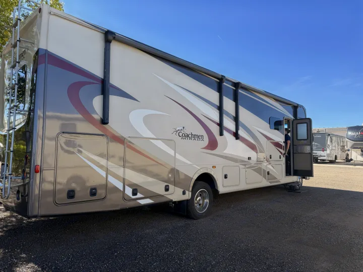 A large, tan Coachmen Mirada rv rental with graphic accents stands on a gravel lot; a person enters through the open door.. Coachmen Mirada 32SS 2019