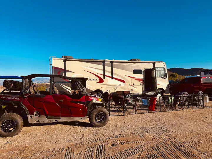 Desert RV rental adventure: a large motorhome and Polaris General UTV parked with a fenced camp area under a clear blue sky.. Coachmen Mirada 32SS 2019