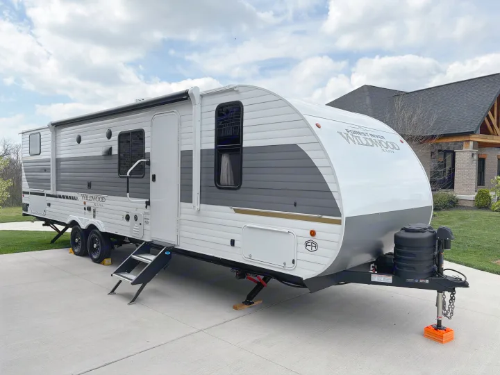 Modern white and grey travel trailer RV rental, parked on a driveway with stabilizer jacks and propane tanks.. Forest River Wildwood X-Lite 273QBXL 2026