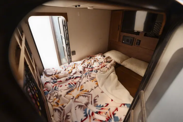 Inside a cozy RV rental bedroom: bed with a colorful blanket, pillows, bright window, TV, and storage.. No Boundaries 10 2020