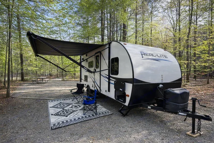 This inviting rv rental travel trailer, with its awning extended over chairs & a rug, is perfectly set up at a wooded spring campsite.. Palomino Real Lite Mini 2017