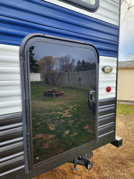 Rear entry door of an RV rental, showcasing a large window reflecting a backyard with a fire pit. Blue, white, & black siding visible.. Forest River Cherokee Wolf Pup 2024