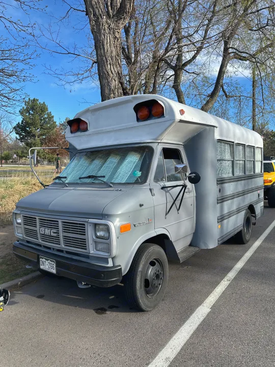 Gray GMC van skoolie RV rental parked under trees on a sunny day. This conversion features a sunshade.. GMC Vandura 1986
