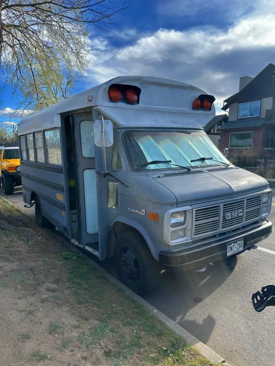 Grey GMC short bus RV rental, a custom conversion, parked with an open door & windshield sunshade.. GMC Vandura 1986