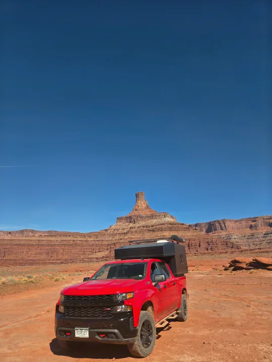 A red pickup truck with an RV camper is parked in a vast desert landscape with striking red rock formations. Ideal for an rv rental adventure!. Chevrolet Silverado 1500 Trail Boss 2020