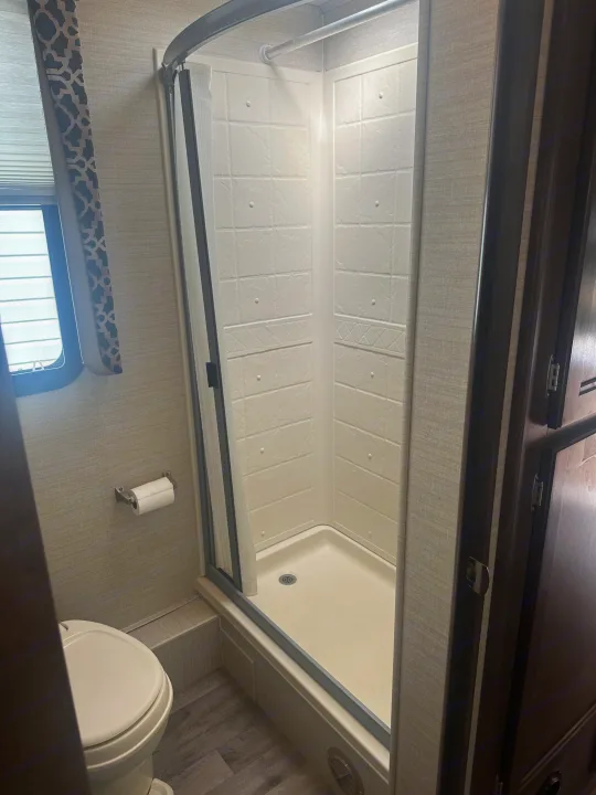 RV rental's compact bathroom: shower stall with textured walls, toilet, and window with patterned curtain.. Jayco Redhawk 2019