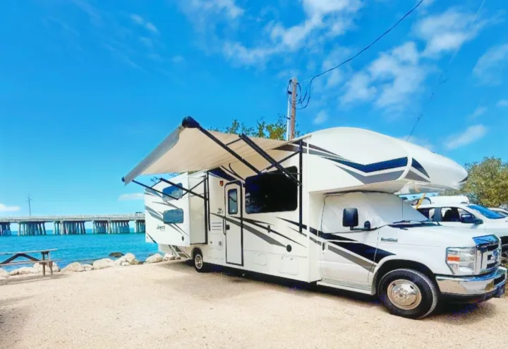Relaxing RV rental parked right by the water with an extended awning, overlooking a distant bridge under a blue sky.. Jayco Redhawk 2019