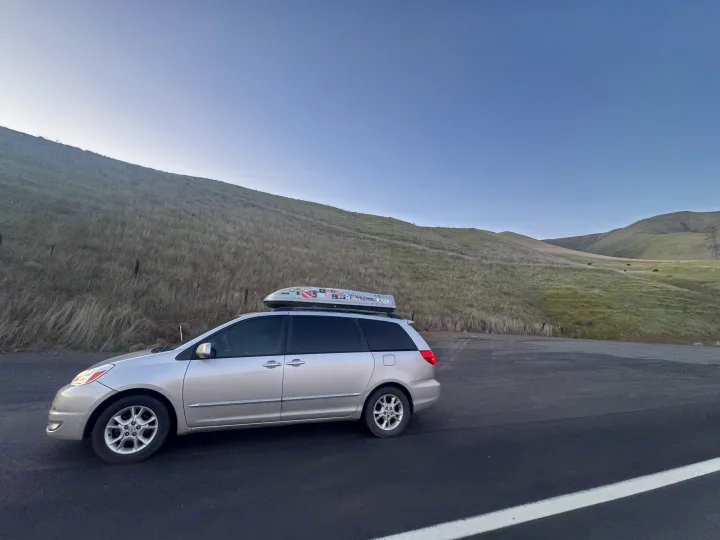 Silver minivan with roof carrier for a road trip, parked by grassy hills. A flexible travel vehicle, perfect for your next RV rental adventure.. Toyota Sienna Limited XLE Camper Van 2004