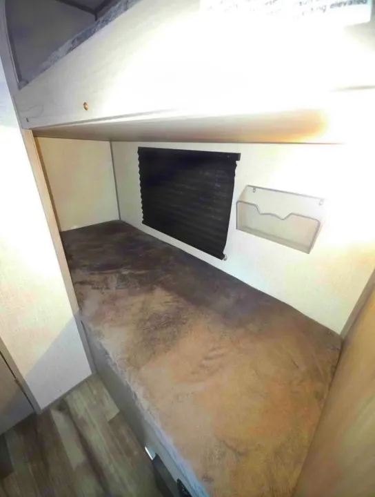 RV rental bunk bed with a soft gray mattress, a dark privacy blind on the window, and a mesh wall organizer.. Dutchmen Coleman Lantern LT 2022