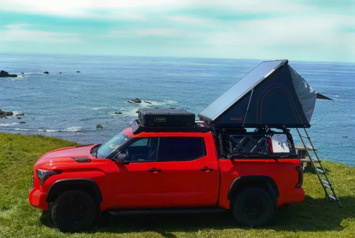 A red overland truck with a deployed rooftop tent and ladder on a grassy ocean cliff. Ideal for an adventurous RV rental trip.. Toyota Landcruiser 2022