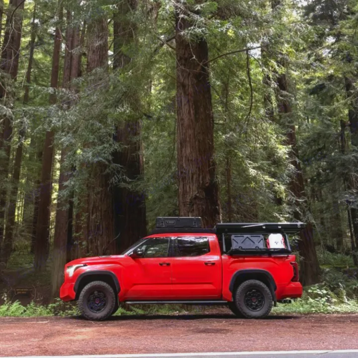A red pickup truck with a camper shell, ideal for an rv rental, is parked by a road amidst towering redwood trees, ready for adventure.. Toyota Landcruiser 2022