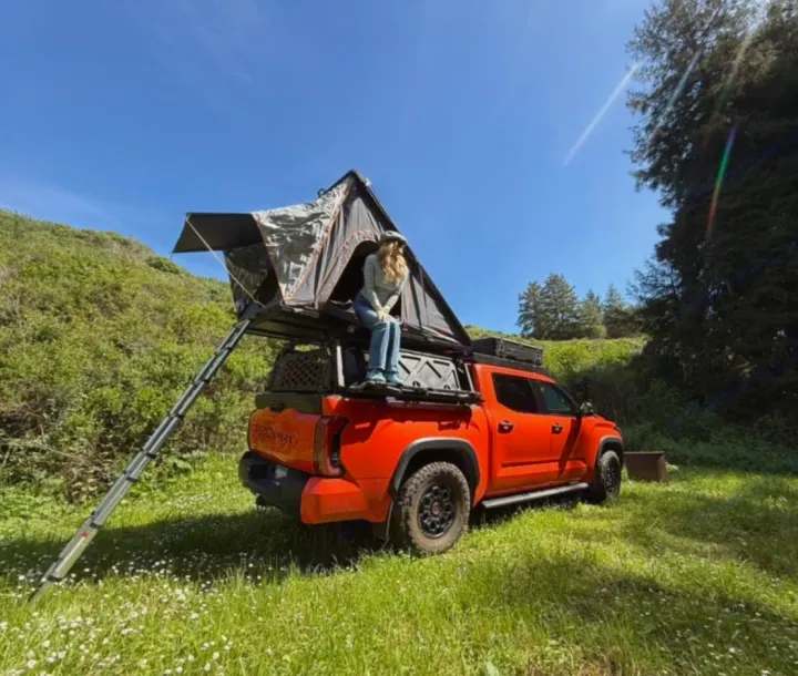 Scenic rv rental: orange overlanding pickup truck with a rooftop tent, a person inside, amidst green hills & blue sky.. Toyota Landcruiser 2022