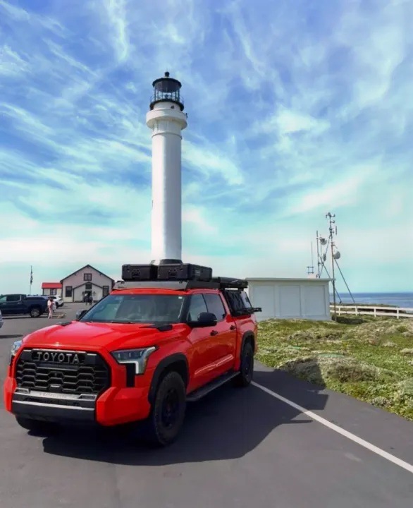 Red Toyota overlanding truck, reminiscent of an rv rental, parked by a tall lighthouse under a cirrus-streaked sky.. Toyota Landcruiser 2022