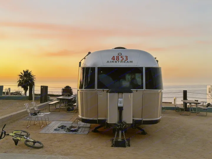 A silver Airstream RV rental sits at a beach campsite during sunset, overlooking the ocean, with a palm tree, picnic area, and bicycle.. Airstream Classic 2017