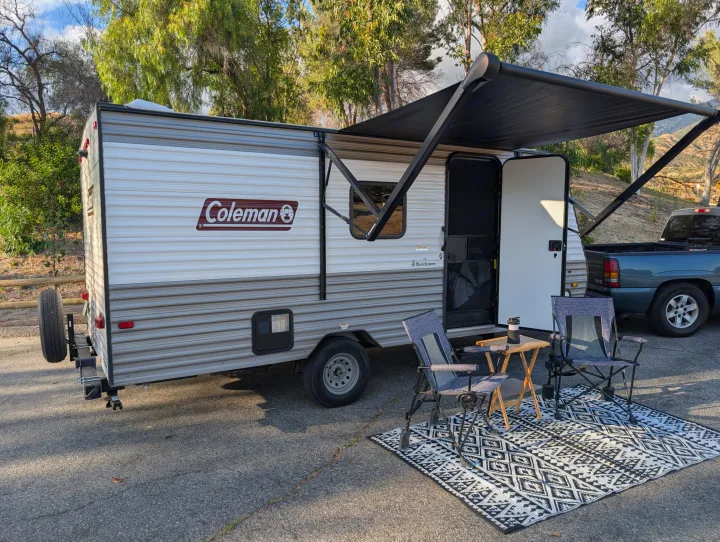 Relaxing with an RV rental: a Coleman travel trailer with an extended awning, two chairs, and a table on a patterned rug.. Dutchmen Coleman Lantern LT 2023