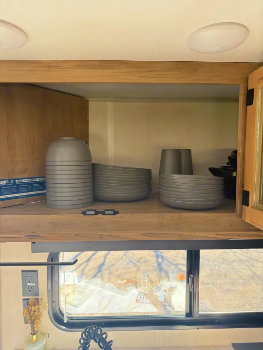An RV rental's overhead kitchen cabinet with stacked gray dishes, cups, and an instruction box. A window shows the outdoors.. Forest River Grey wolf 2026