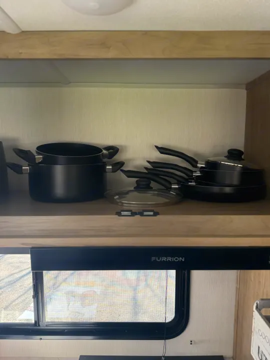 RV rental kitchen showing black pots & pans neatly stored in an overhead cabinet, with a window and "Furrion" range hood below.. Forest River Grey wolf 2026