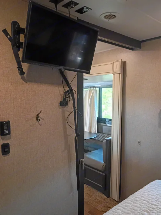 Inside an RV rental: a wall-mounted TV is secured with a strap, with a thermostat and hook nearby. A dinette is seen through the doorway.. Jayco Eagle HT 2020
