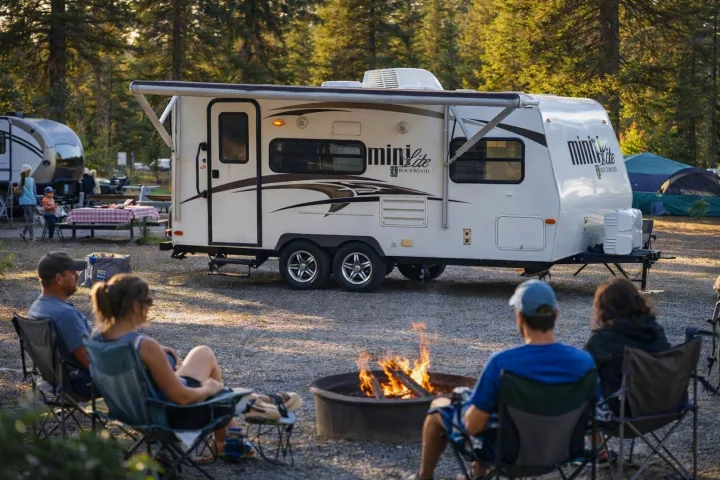 Enjoying a campfire at an RV rental campsite. People relax around the fire, with a white Mini Lite travel trailer nearby.. Forest River Rockwood Mini Lite 2014